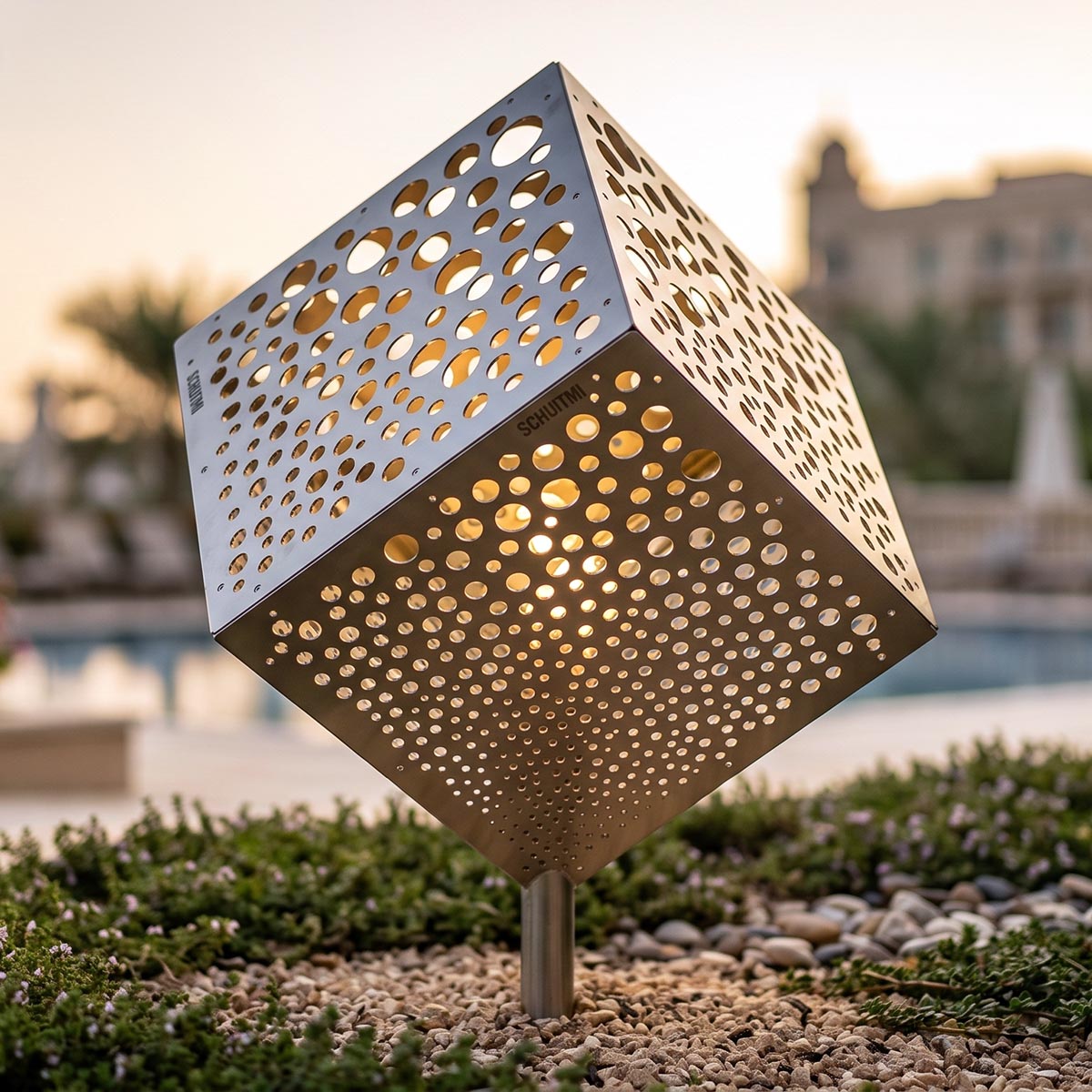 A modern geometric stainless steel cube solar light fixture with intricate laser-cut circular patterns, mounted on a single metal post. The light is turned on, casting a warm golden glow through the perforations.