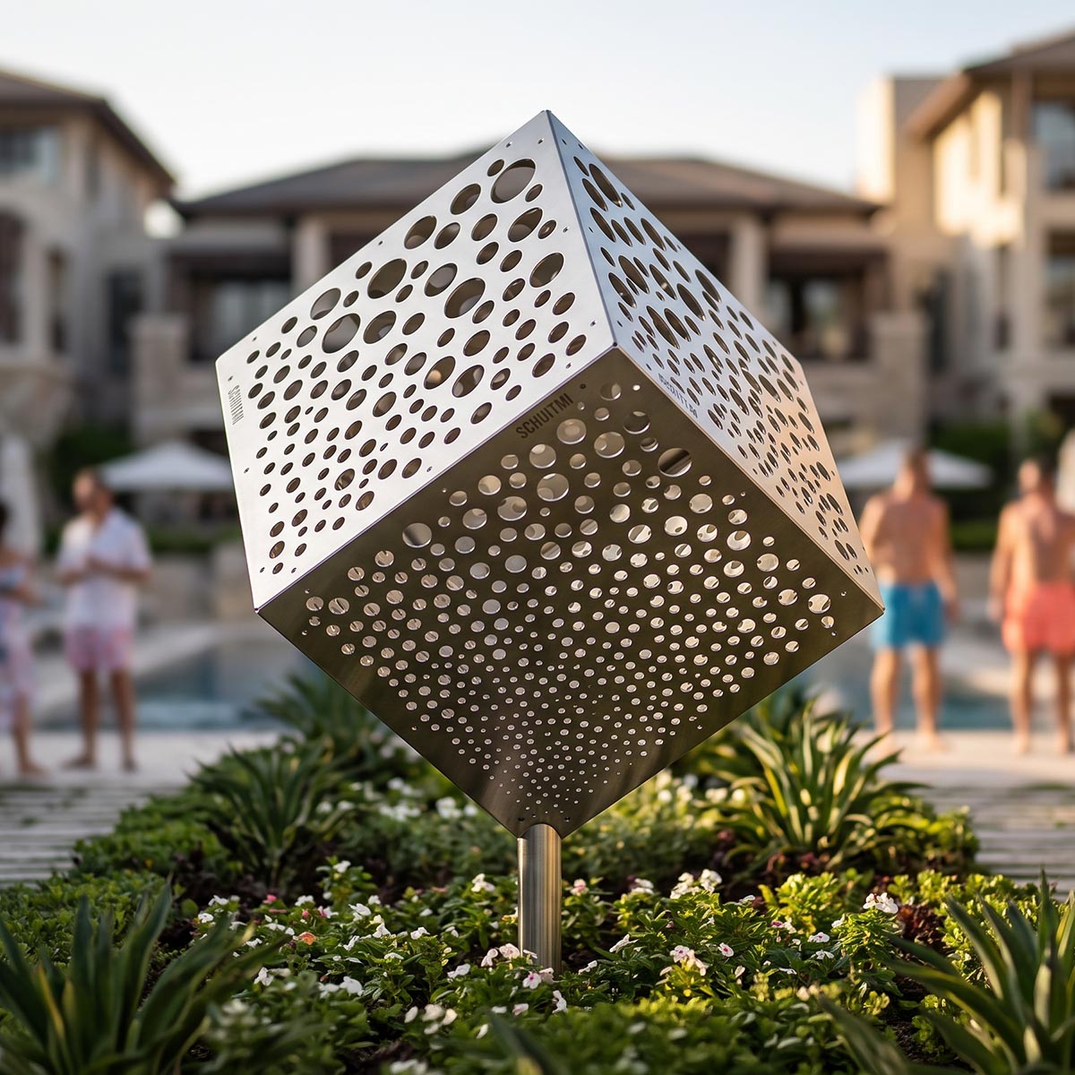 A modern perforated stainless steel cube garden sculpture on a slender post, set in a landscaped garden with blurred people and poolside buildings in the background.